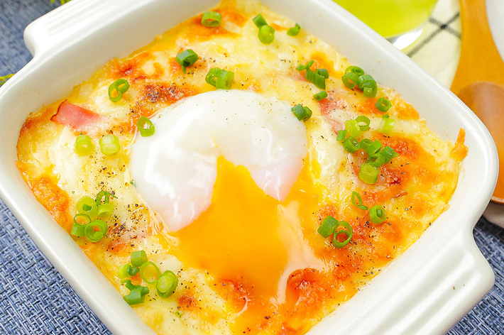 熱々のグラタンの上に半熟卵が乗り、刻みネギが散らされている料理。背景に黄色のドリンクが映っている。