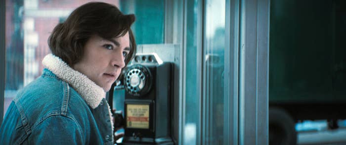 Michael Gandolfini in a jean jacket with shearling collar, looks through a phone booth in a film scene