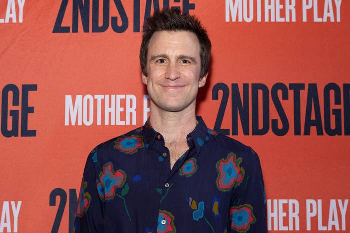 Gavin Creel stands in front of a red backdrop featuring the text &quot;2NDSTAGE&quot; and &quot;MOTHER PLAY.&quot; He is wearing a dark button-up shirt with a floral pattern