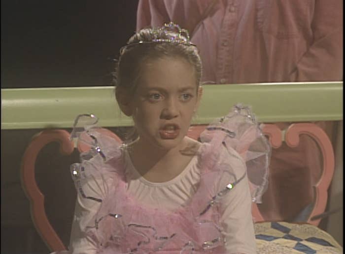A young Laurie dressed as a ballerina with a tiara is sitting and talking. An adult stands behind her
