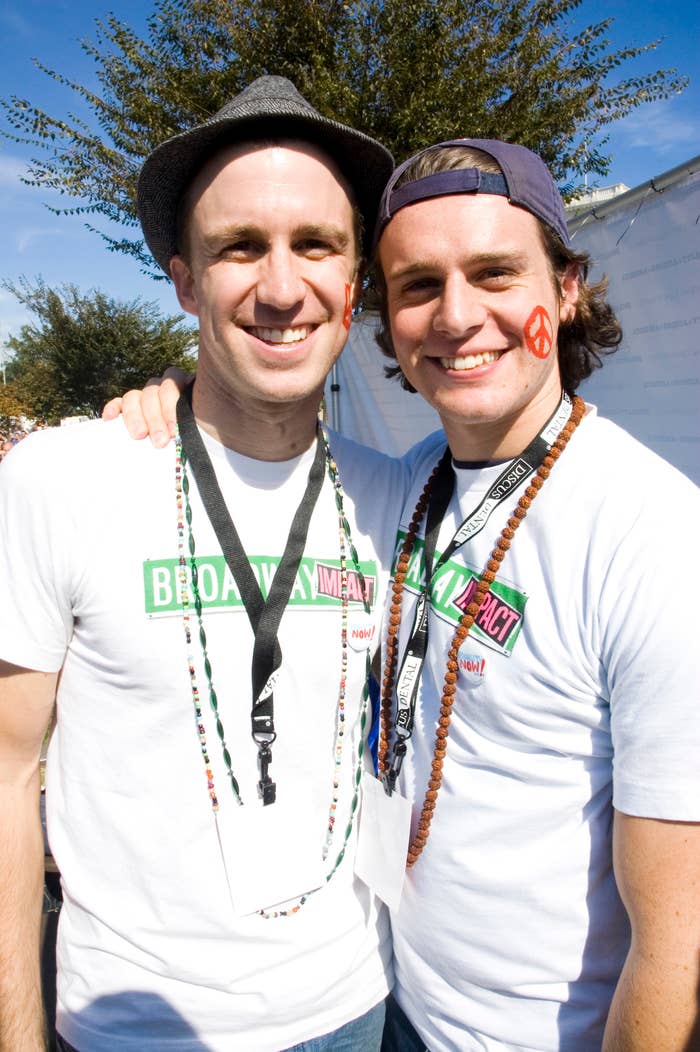 Gavin Creel and Jonathan Groff smile together at an outdoor event. Both wear Broadway Impact t-shirts and necklaces.