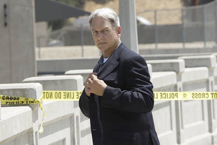 Mark Harmon wearing a suit, standing near police tape at an outdoor crime scene