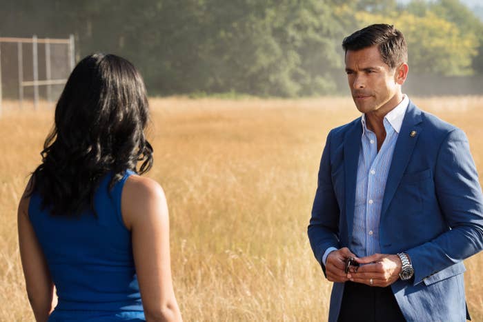 Camila Mendes and Mark Consuelos in a field scene, Mendes facing away in a sleeveless dress, and Consuelos in a blazer and shirt, holding an object