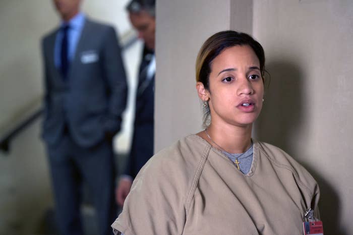 Dascha Polanco, in a prison uniform, appears concerned in a scene from a TV show, with two blurred men in suits in the background