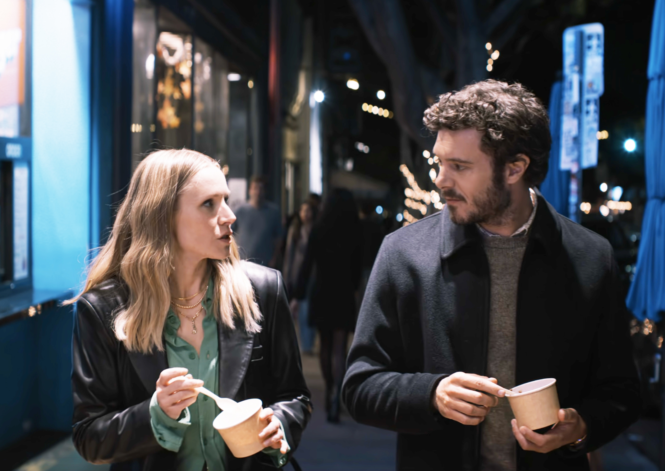 Kristen Bell and Adam Brody walk at night while eating ice cream, engaged in conversation, on a street decorated with lights