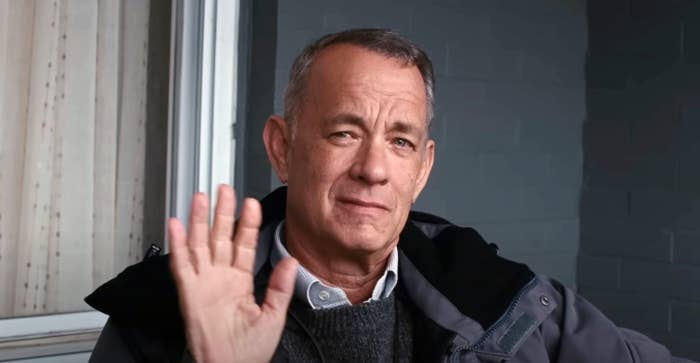 Tom Hanks waves with a slight smile, wearing a dark jacket and sweater. He is seated indoors next to a window with blinds