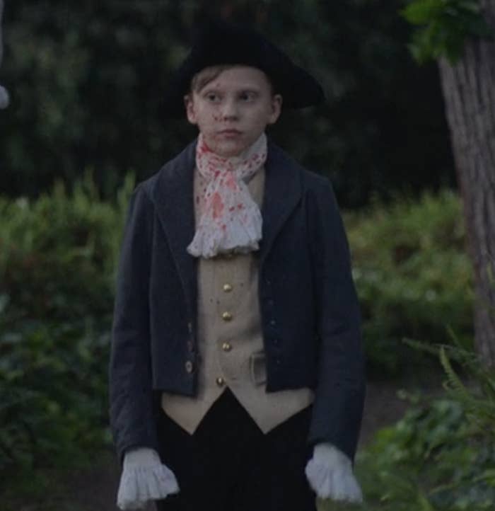 A young Stede in historical attire with a tri-corner hat, gloves, a dark coat, and a blood-splattered  neck scarf, stands outdoors among greenery