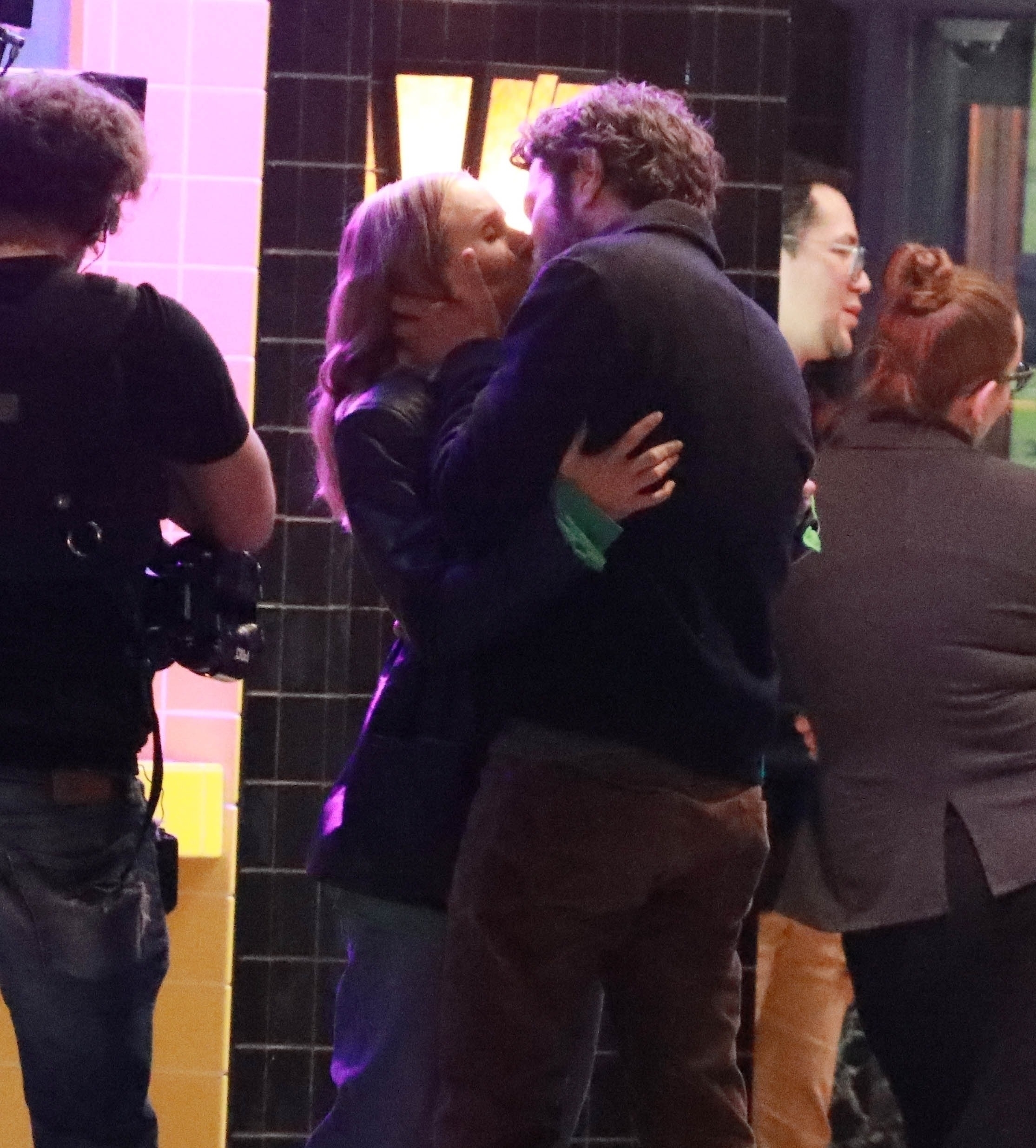 Kristen Bell and Adam Brody share a kiss on a TV or movie set, surrounded by crew members. Kristen wears a jacket; Adam is in a sweater