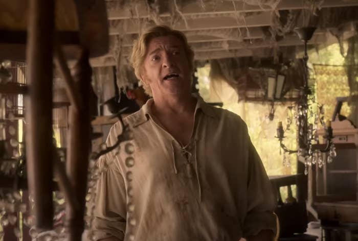 Rhys Darby dressed as Stede Bonnet in a rustic setting from &quot;Our Flag Means Death,&quot; wearing a light, old-fashioned shirt