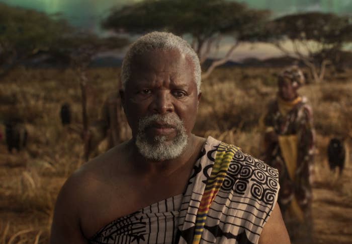 John Kani wearing traditional attire in a savanna landscape