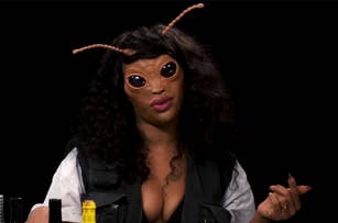 A person wearing an elaborate insect-themed mask with antennae and large black eyes is talking or posing for the camera
