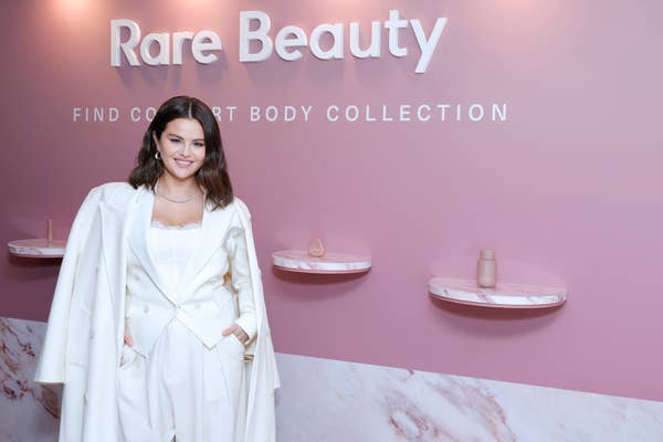 Selena Gomez at a Rare Beauty event, wearing a stylish outfit under a white coat, standing in front of a display for the &quot;Find Comfort Body Collection.&quot;