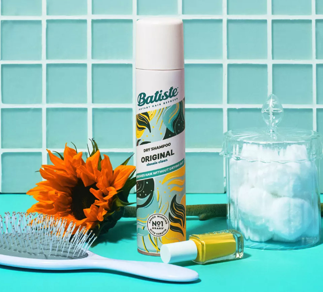 Batiste Original Dry Shampoo on a tiled background, alongside a hairbrush, a sunflower, a nail polish bottle, and a jar filled with cotton balls