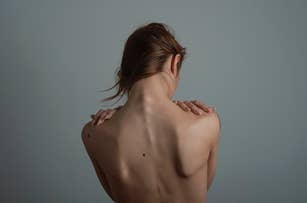 A person with light skin, seen from behind, covering their shoulders with their hands. The focus is on the back and shoulders without any visible clothing