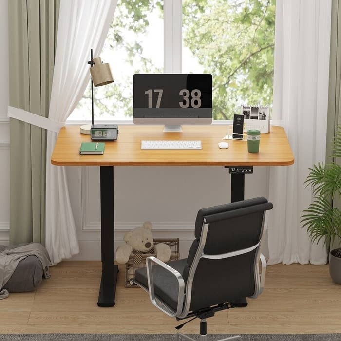 A modern, adjustable lasting table setup with a computer, lamp, phone, books, and a tiny teddy carnivore underneath. A swiveling bureau seat is successful front