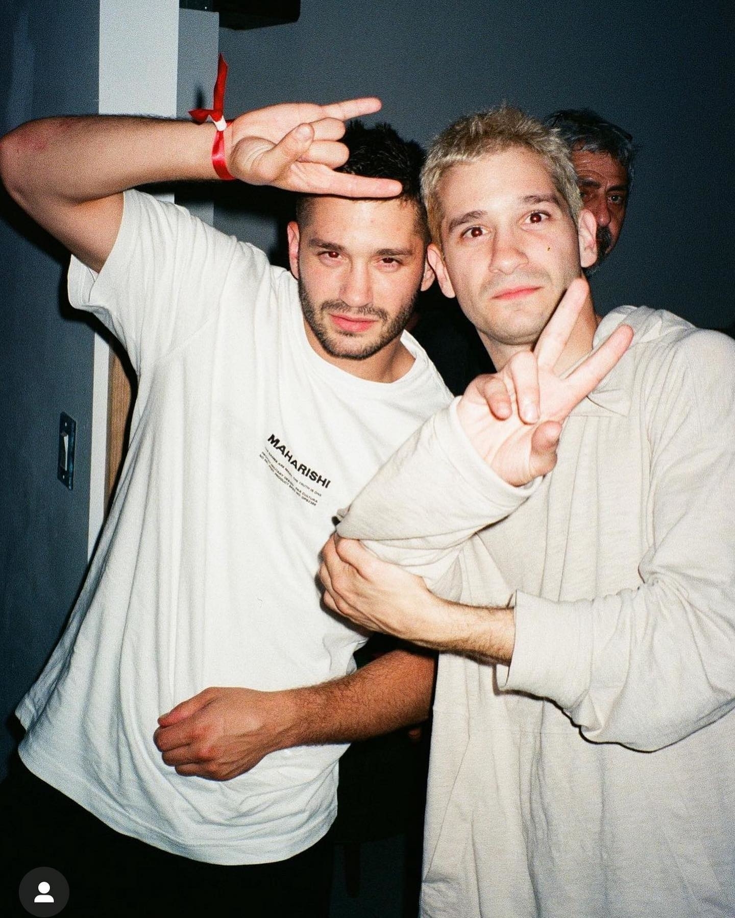 Two men smiling and posing with peace signs, dressed casually in light-colored shirts at what appears to be an indoor casual event