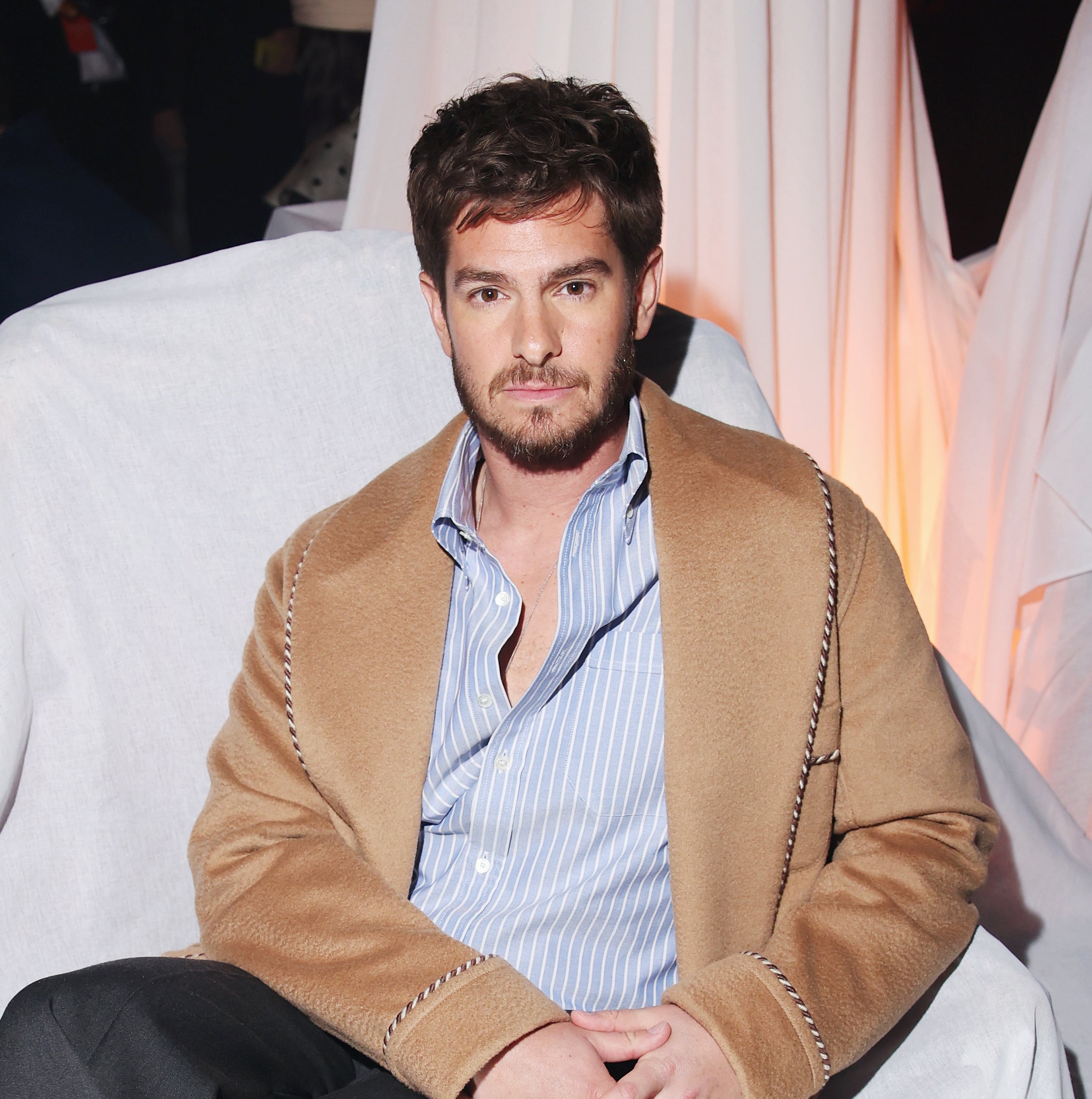 Andrew Garfield seated in a stylish outfit with a blazer over a shirt, tailored trousers, and loafers