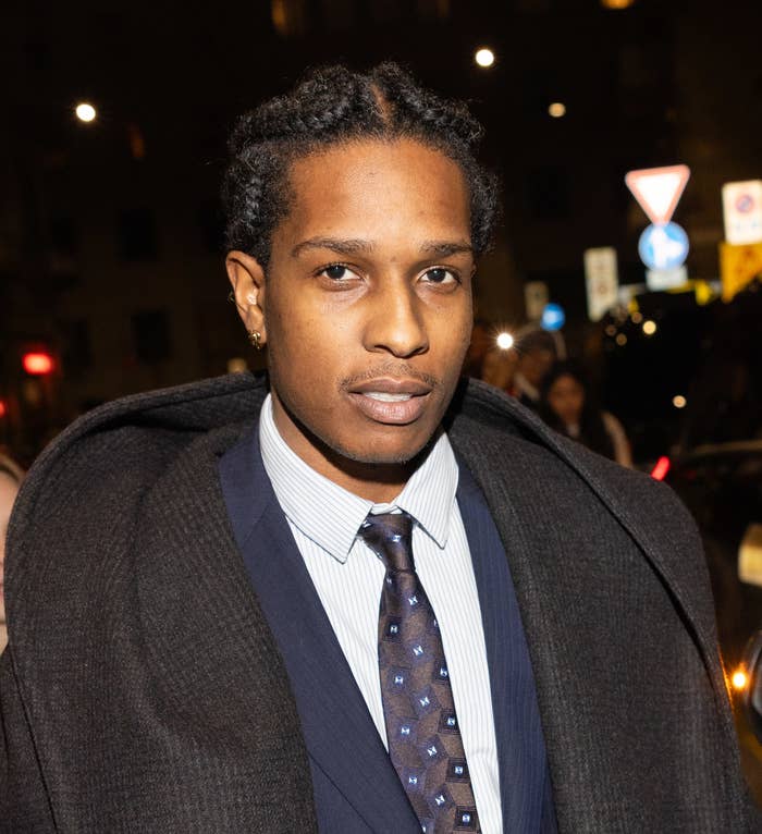 A$AP Rocky in a suit and tie with a coat draped over their shoulders attends an event
