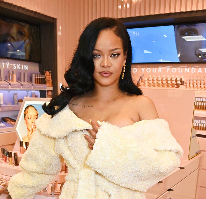 Rihanna wearing an off-shoulder fluffy coat and heels stands in a Fenty Beauty store filled with cosmetics displays