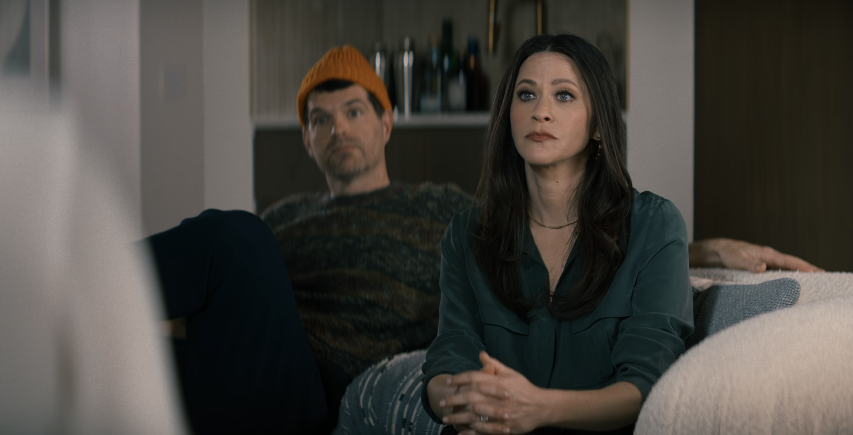 Jackie Tohn sits upright on a coach with a serious expression and her hands crossed