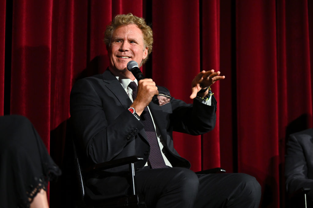 A person in a suit sits on stage speaking into a microphone with a red curtain background