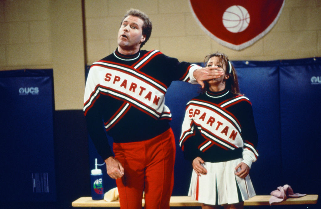 Two adults, one in red pants and the other in a skirt, wear matching &quot;Spartan&quot; cheerleader outfits in a comedic scene