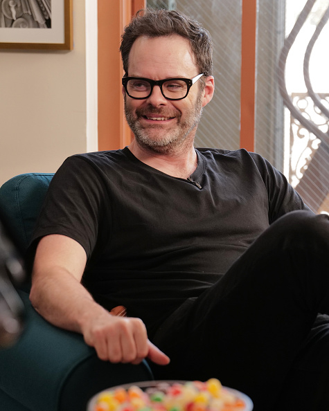 A man in casual black attire sits on a blue chair, smiling, with a bowl of candy visible in the foreground