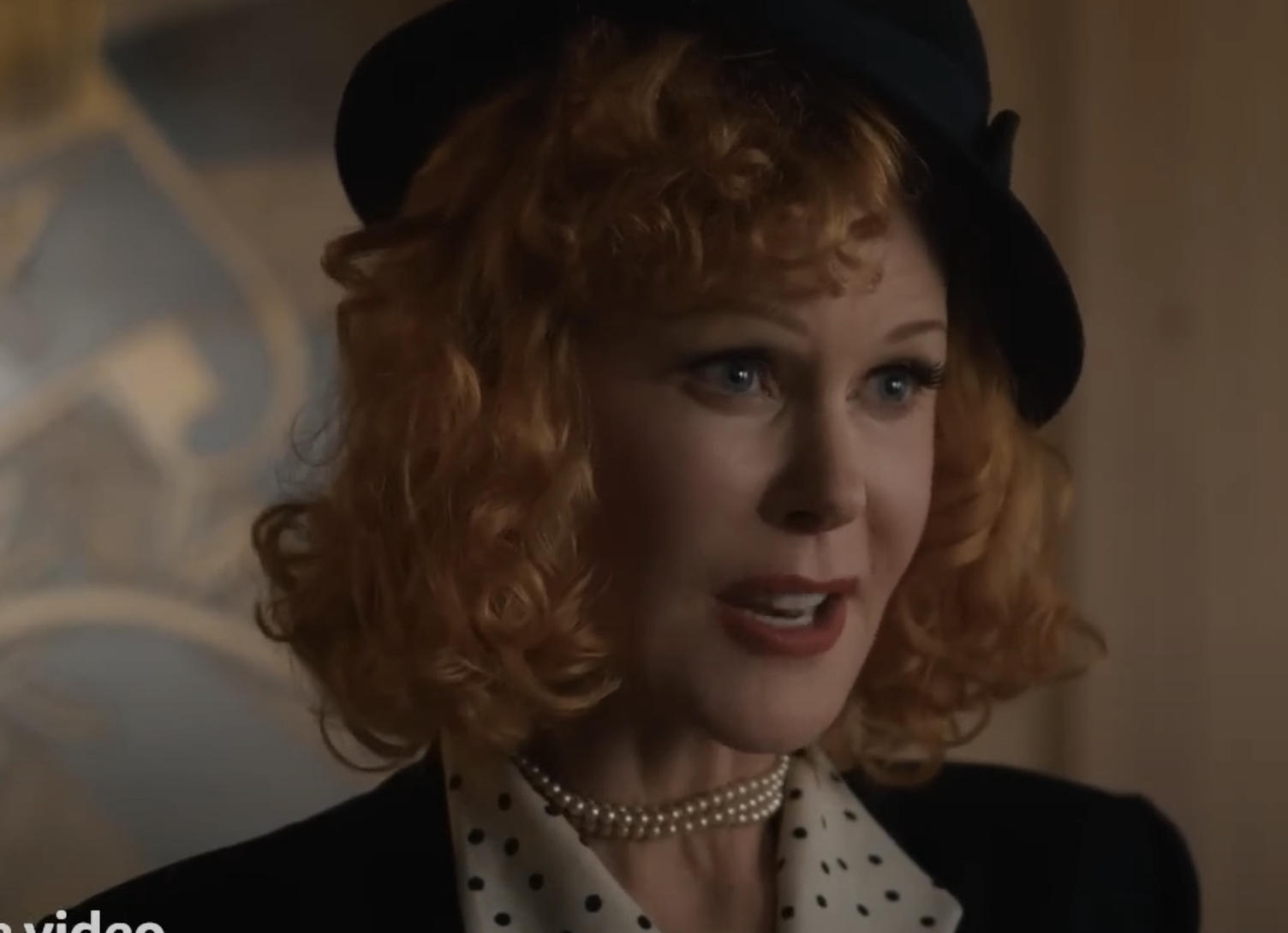 I don't know who is in the image. A woman with curly hair wearing a hat, polka dot shirt, pearl necklace, and jacket is shown speaking