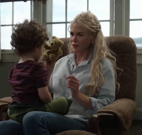 A woman with long blonde hair is sitting in a chair, holding a teddy bear with a child standing in front of her