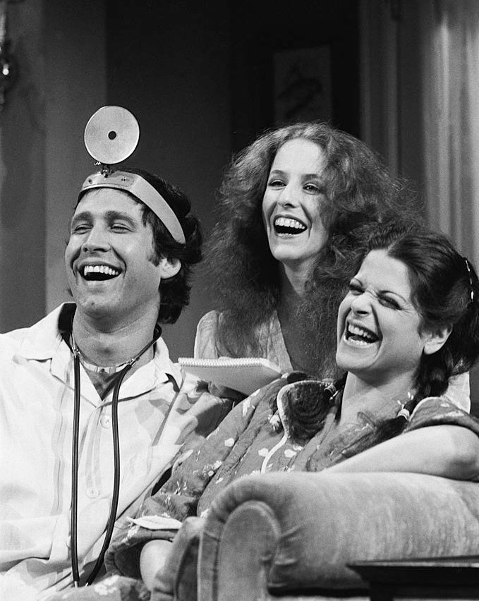 Three people laughing, one wearing a stethoscope and head mirror, sitting on a couch