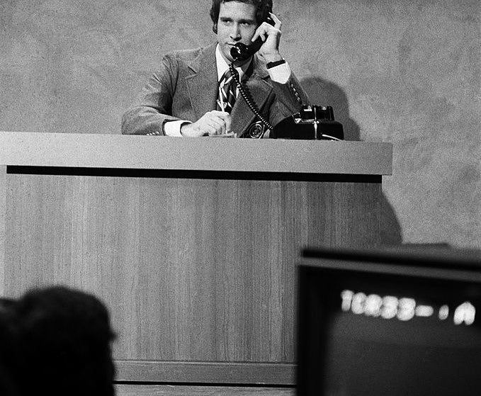 A person in a suit sits at a news desk labeled &quot;Weekend Update,&quot; speaking on the phone. A TV camera is visible in the foreground