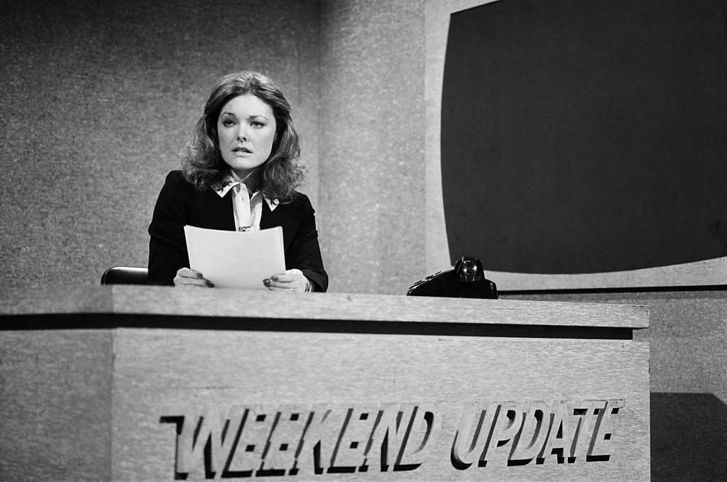 A woman sits at a news desk for &quot;Weekend Update&quot; holding papers, wearing a suit with a collar