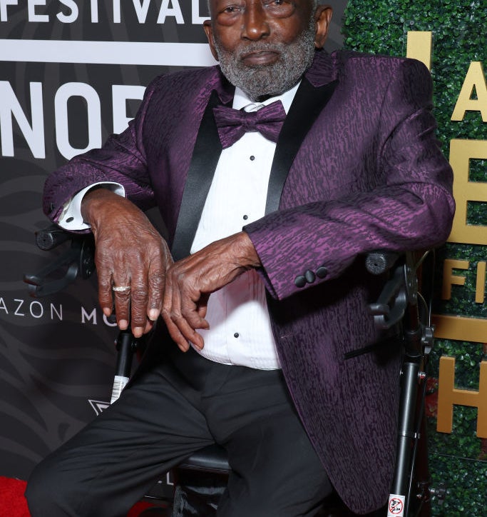 An elderly man in a patterned suit with a black bow tie is sitting in a wheelchair on the red carpet at an event