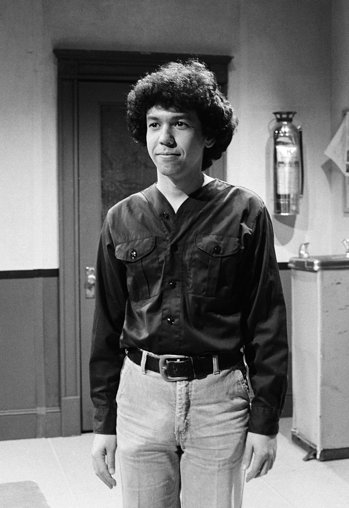 A person with curly hair stands in a room, wearing a long-sleeved shirt and jeans