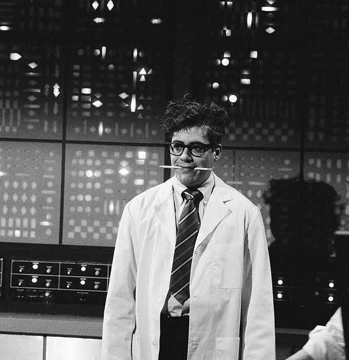 A person in a lab coat and tie stands in front of a retro control panel, appearing to mimic a scientist, with a humorous expression