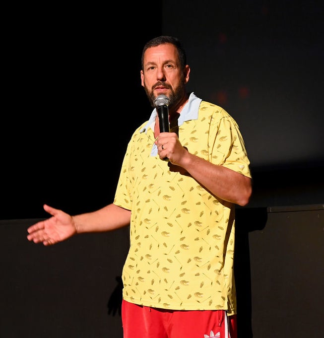 A person speaking on stage, wearing a casual yellow shirt with a pattern, and red pants, holding a microphone