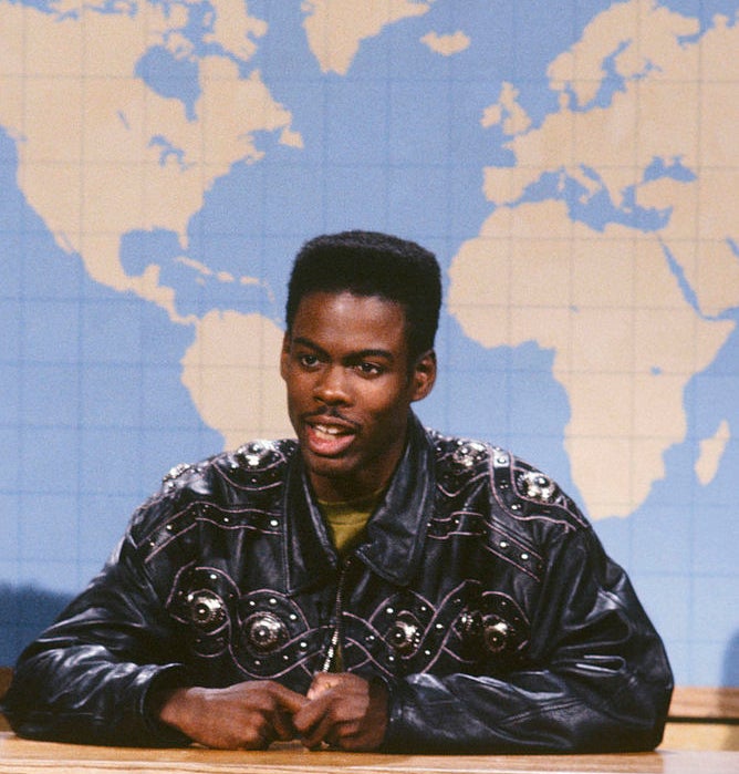 A person sits at a news desk on a set with a world map backdrop, wearing a studded leather jacket