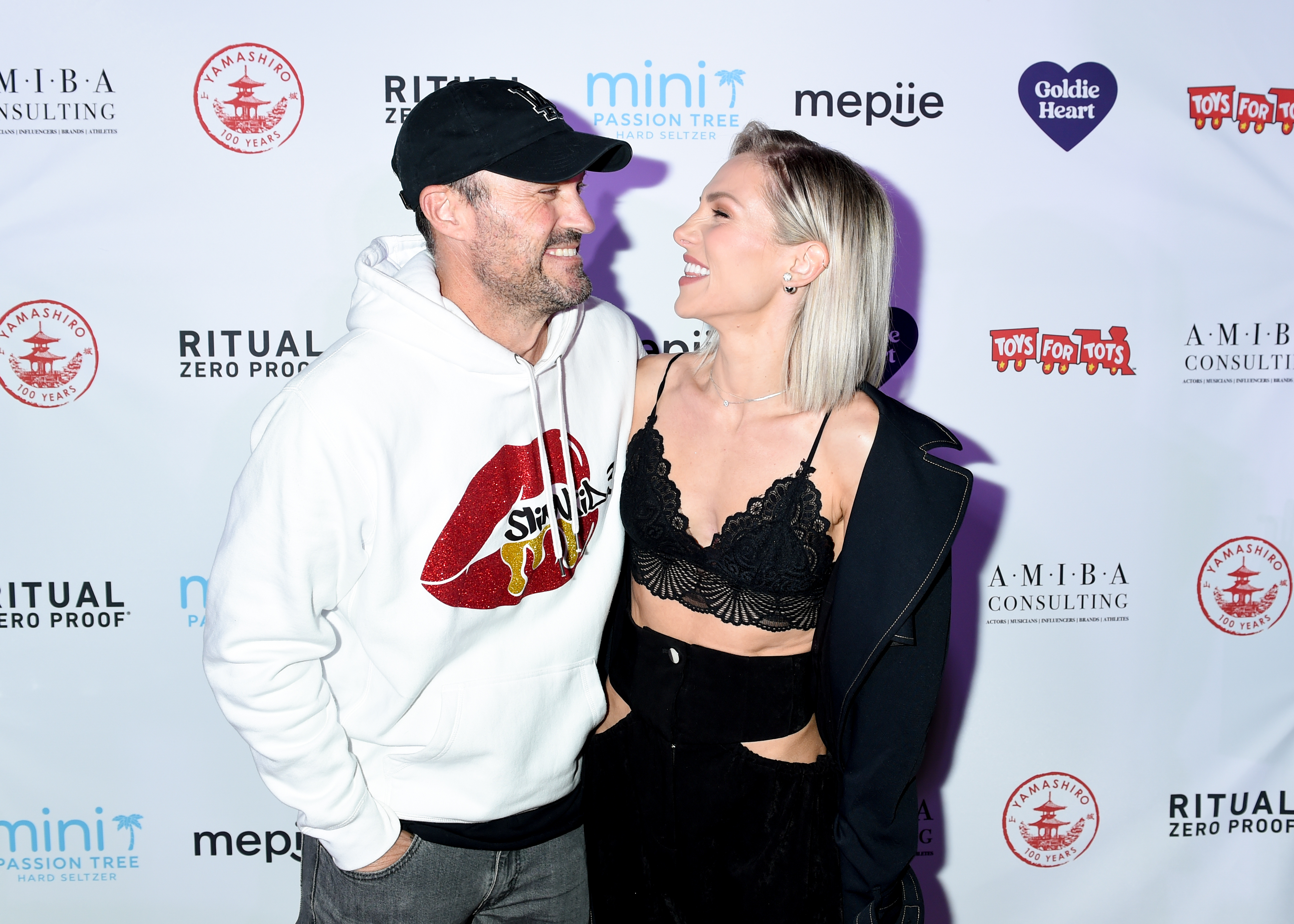 Brian Austin Green and Sharna Burgess smiling at each other on a red carpet. The woman wears a black lace top and the man wears a hoodie with a graphic