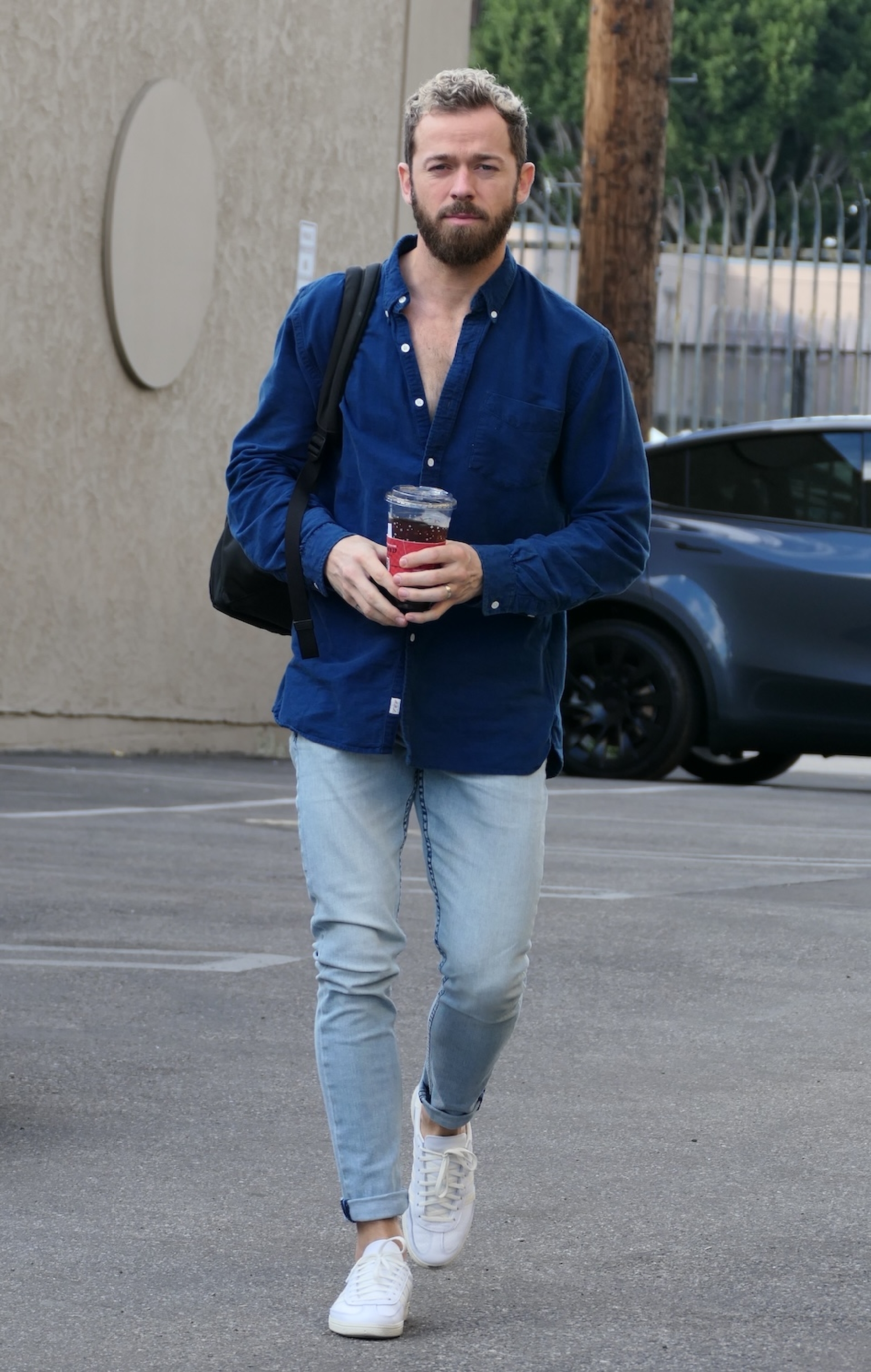 Artem Chigvintsev with a beard wearing a blue shirt, light jeans, and white sneakers, holding a drink and bag, walks in a parking area