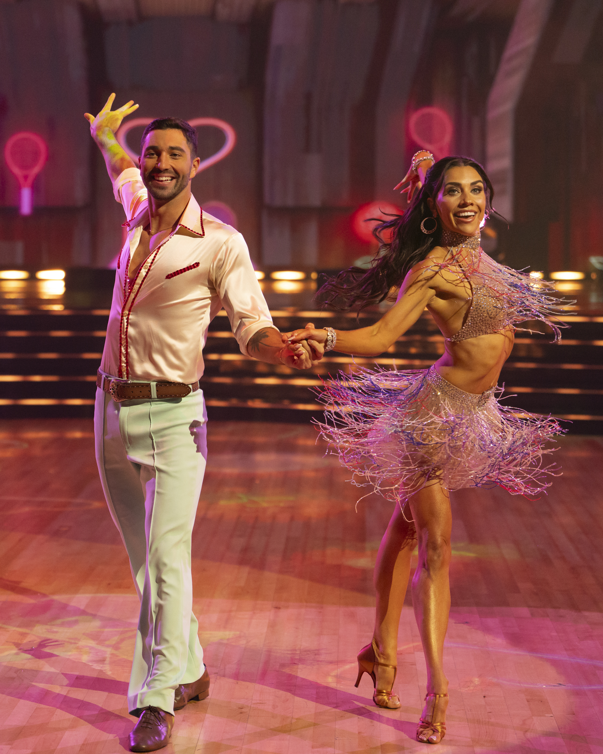 A man and woman dance energetically on a set resembling a ballroom show, with the woman in a shimmering fringe outfit and the man in a stylish shirt and pants