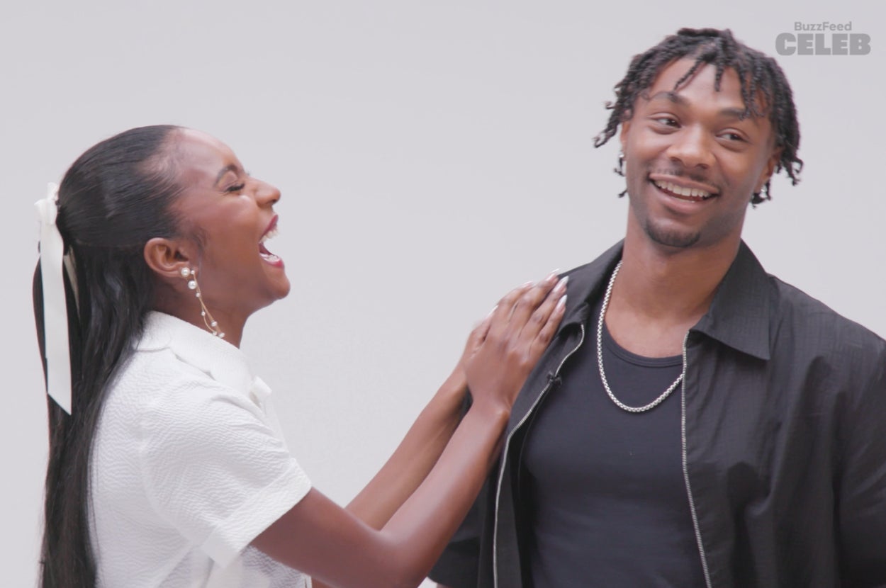 Two people smiling and laughing together. One is wearing a white shirt with their hair tied back, and the other is in a black jacket with layered necklaces