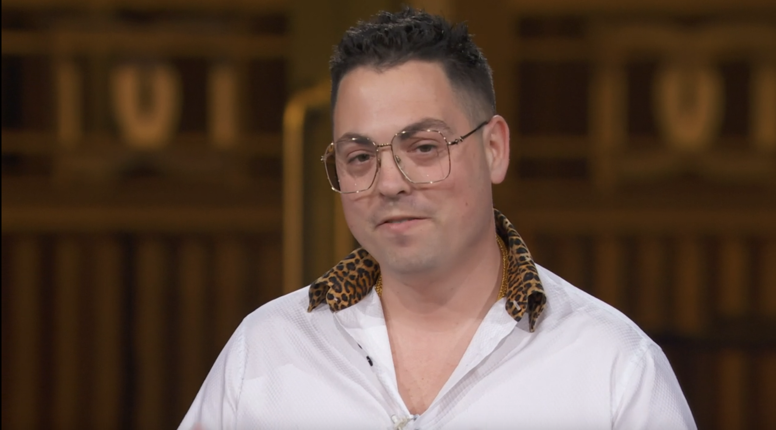 A person in a white shirt with a leopard print collar and glasses stands against a blurred background