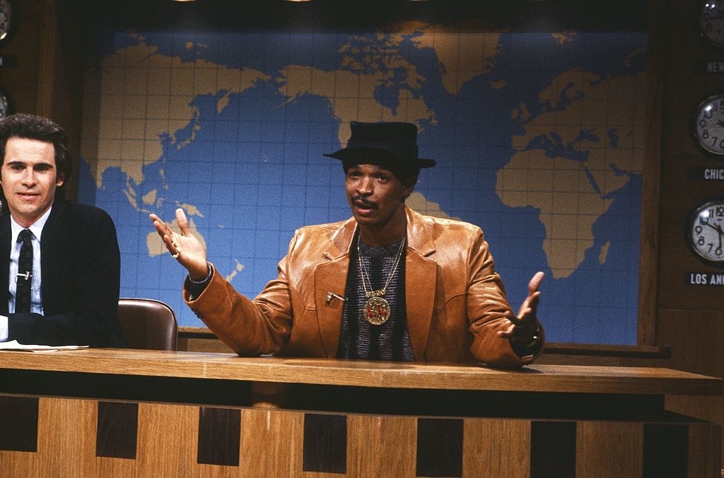 Two people sitting at a news desk; one wears a suit, the other a leather jacket and hat, gesturing as they speak, with world map background