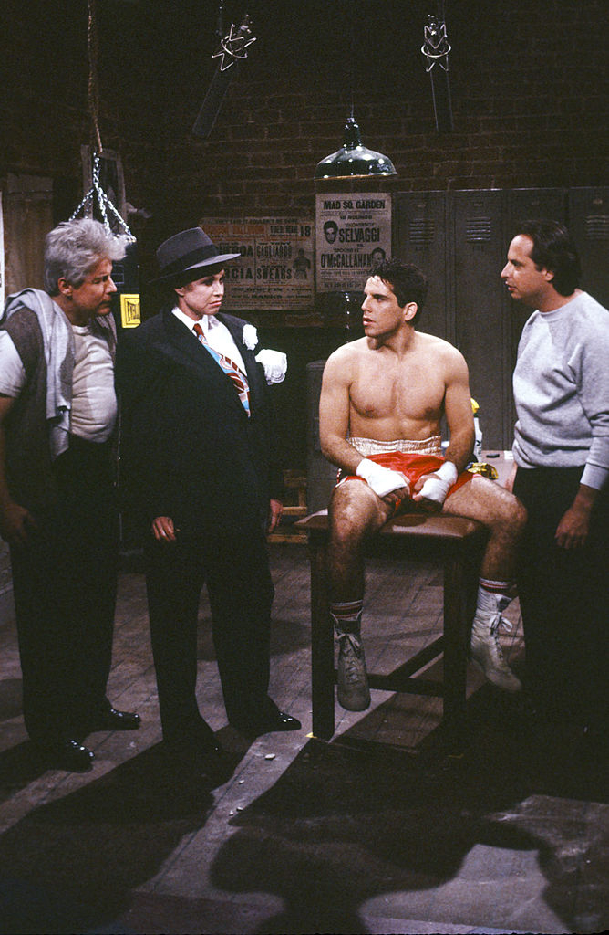 I don’t know who these individuals are. The image shows four people in a boxing gym, with one person in boxing gear sitting and others standing around