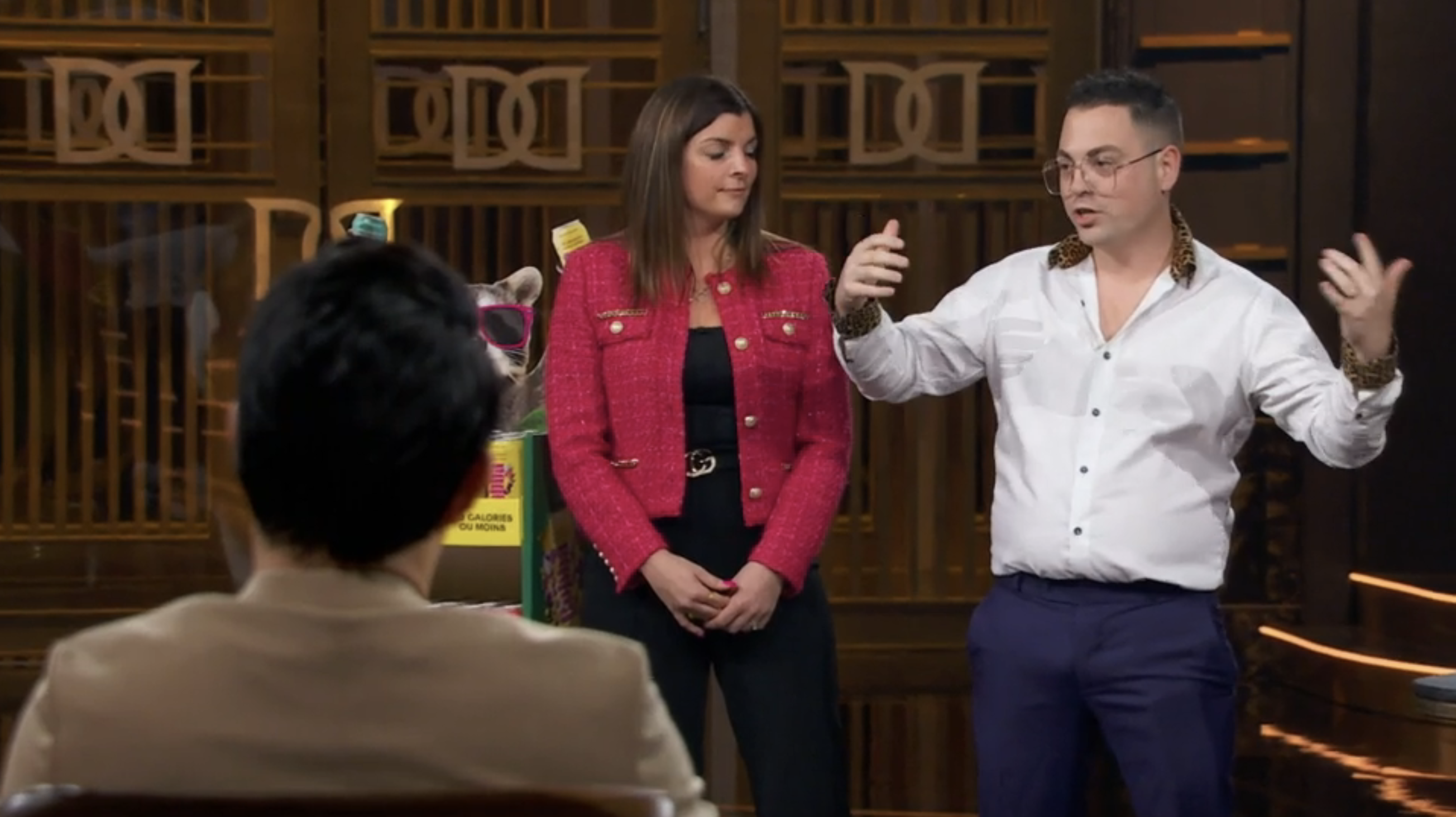Two people present in a business setting, one wearing a red jacket, the other a white shirt with patterned cuffs. Audience partially visible