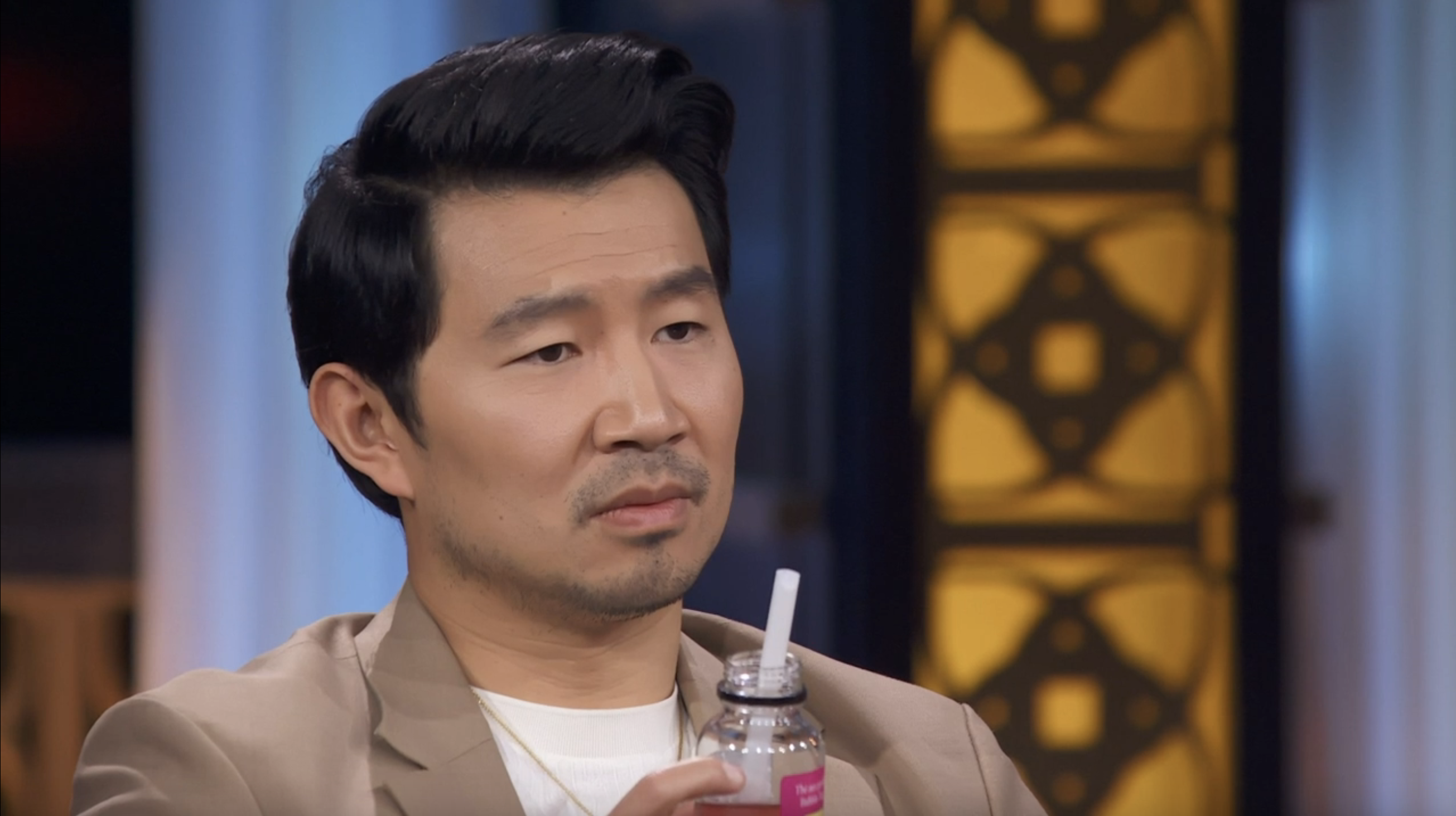 A person in a tan jacket holds a drink with a straw during an interview