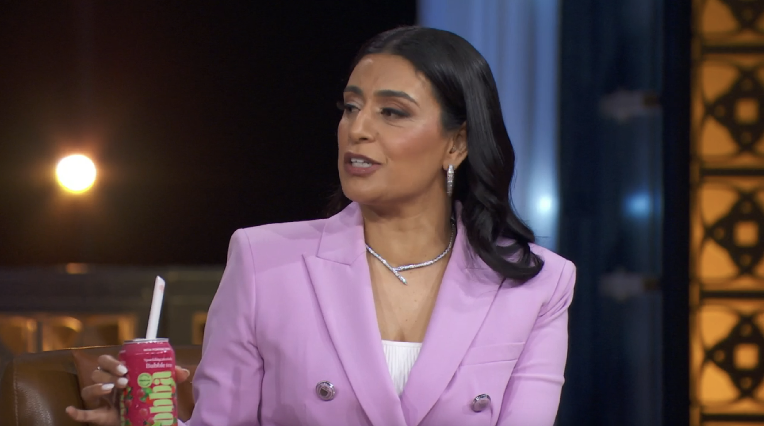 A person in a stylish blazer holds a bubble tea cup during an interview or talk show appearance