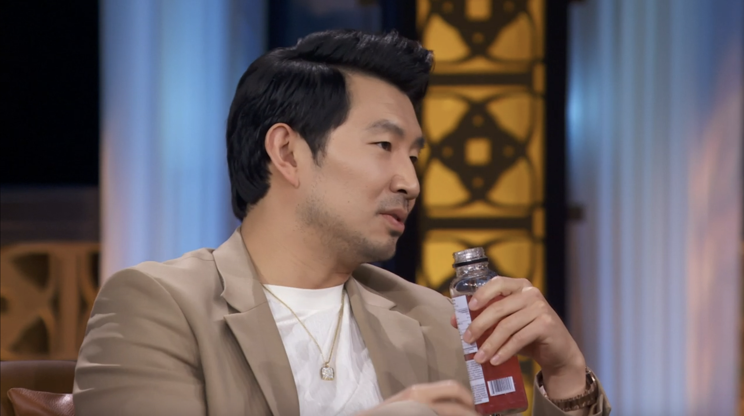 Man in beige suit holding a drink while talking on a TV show set