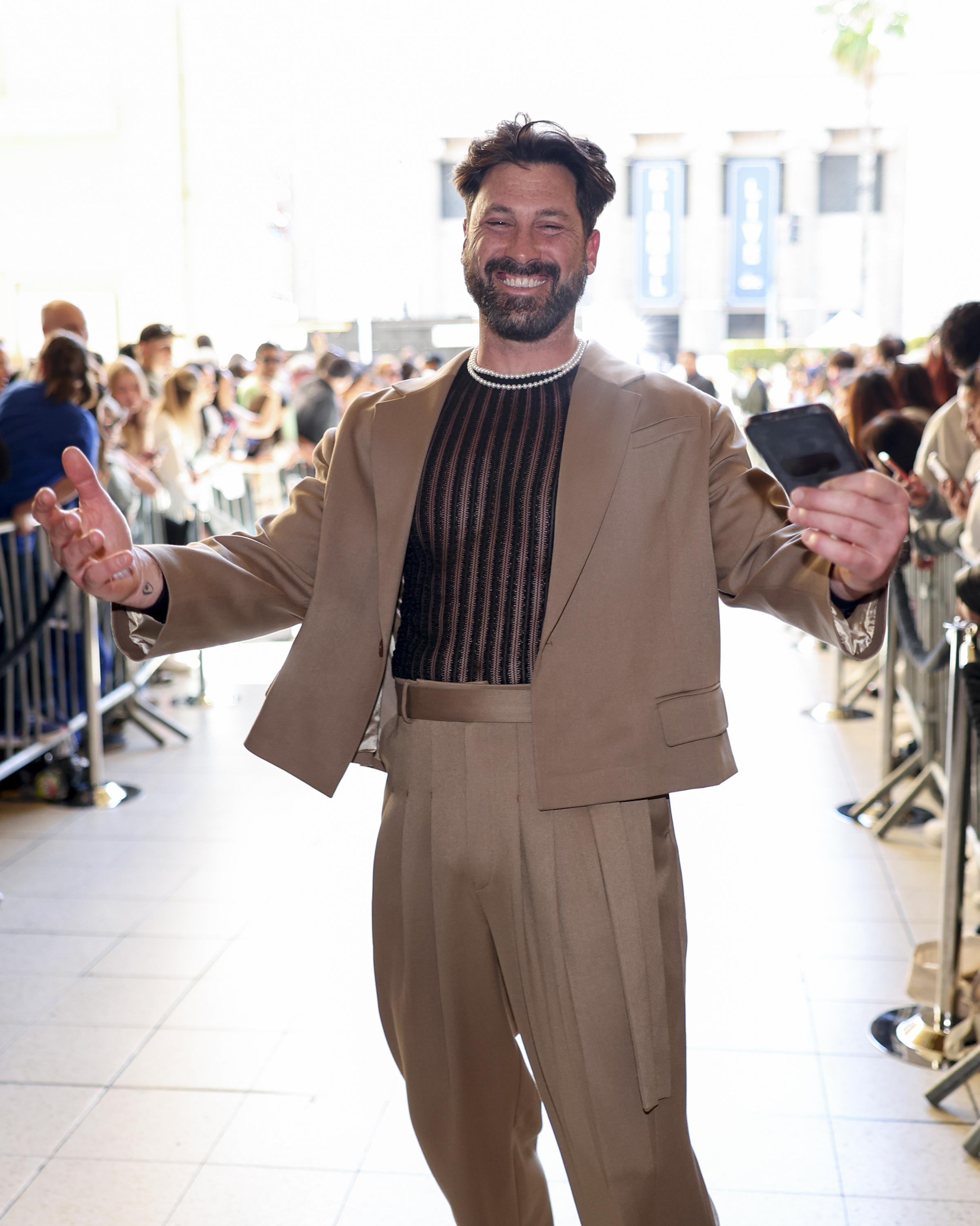 A person in a stylish tan suit with a black sheer top poses for the camera, holding a phone and smiling at a public event