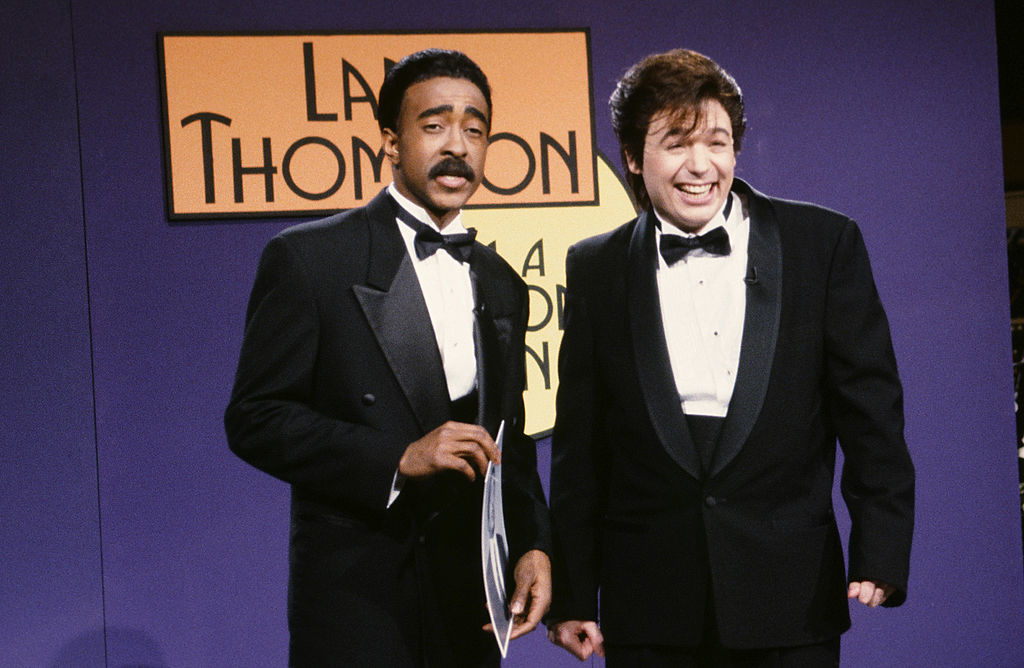 Two men in tuxedos stand on a stage in front of a sign with text